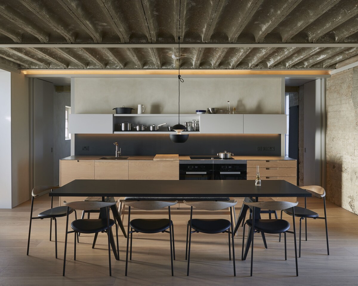 An open-plan kitchen area features a modern design with a light wood cabinetry and sleek countertops. A dining table, seating eight, stands in the center, surrounded by black chairs. The space is illuminated by a pendant light and has ample storage with visible kitchenware displayed.