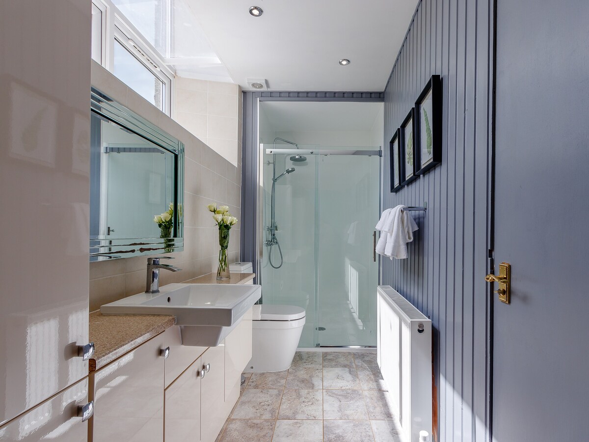 A modern bathroom features a spacious vanity unit with a minimalist sink, surrounded by light-colored tiles. A glass shower enclosure stands alongside a toilet, while a vase of fresh flowers adds a touch of nature. Natural light floods the room through high windows.