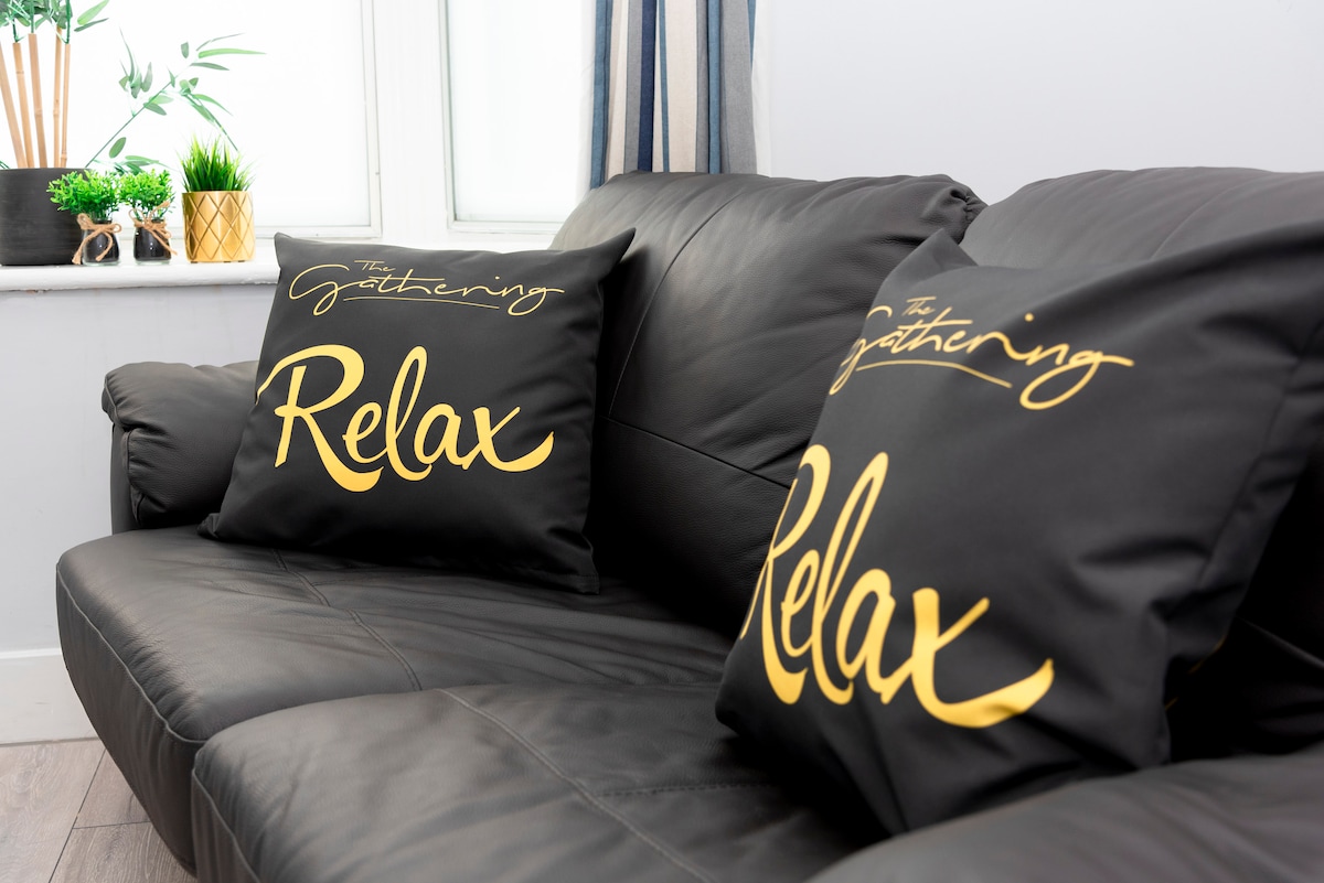 A dark leather sofa features two decorative pillows with the words 'The Gathering' and 'Relax' printed in gold. Natural light filters through a nearby window, enhancing the cozy seating area, which includes a small plant on the windowsill.