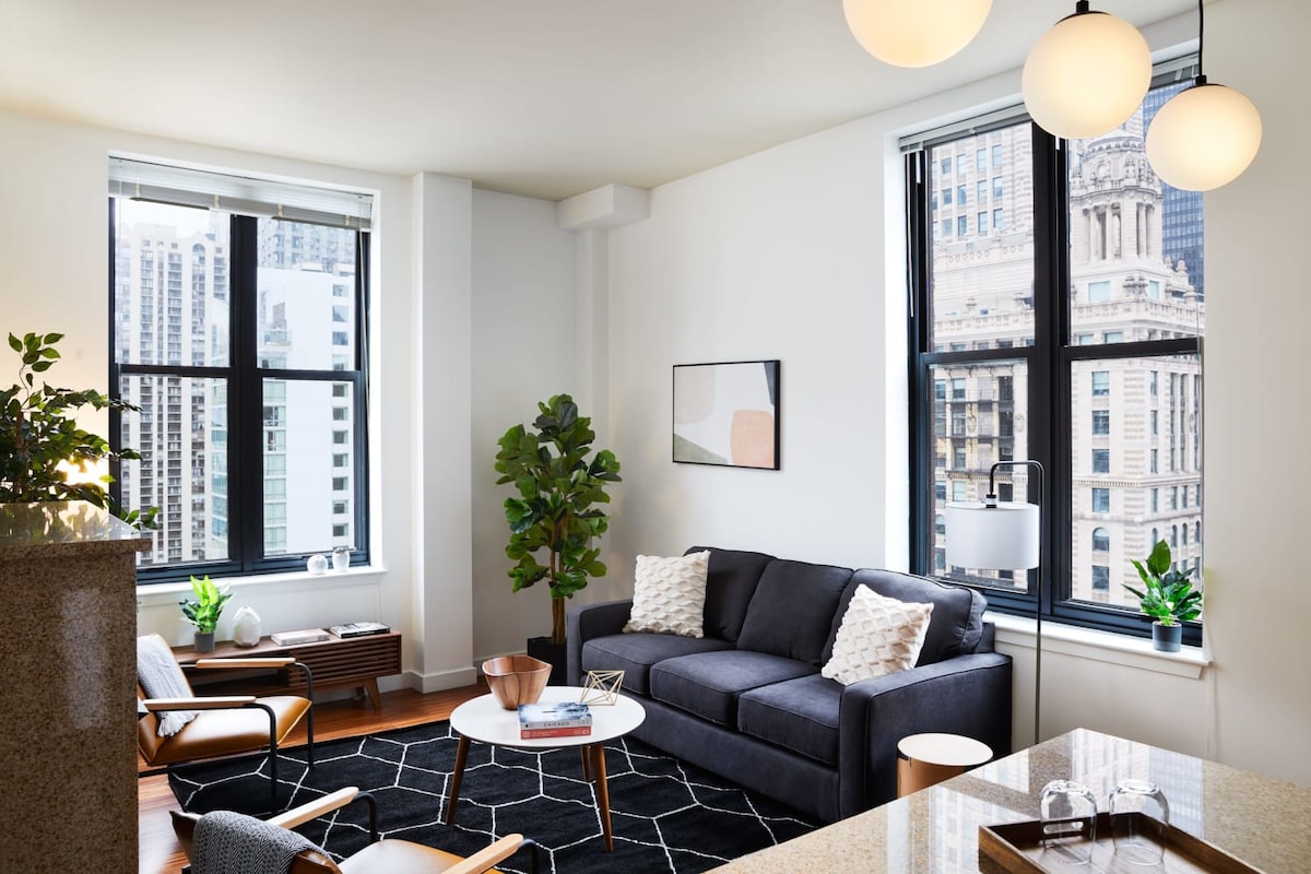 A modern living room features a comfortable dark sofa adorned with decorative cushions. A geometric area rug lies on the hardwood floor, complemented by a round coffee table. Large windows provide an abundance of natural light, showcasing the cityscape outside.