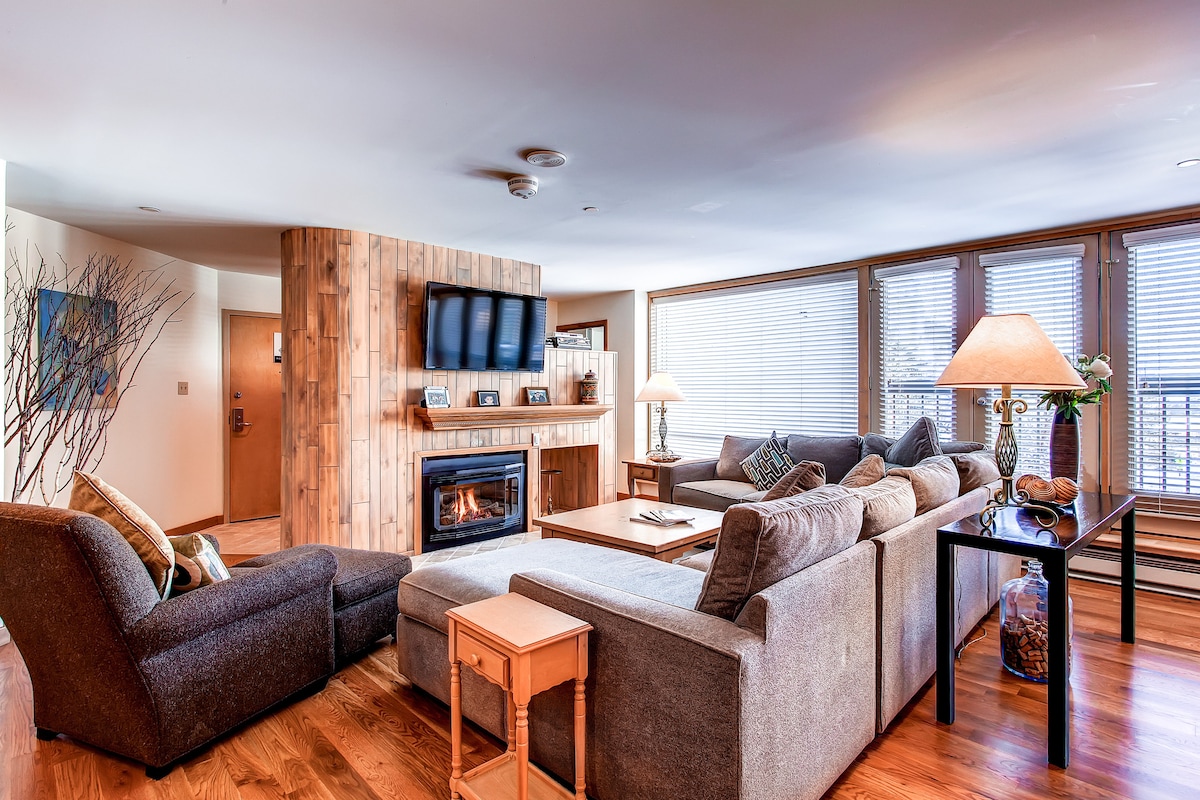 The living area features a cozy sectional sofa, a comfortable armchair, and a coffee table. A television is mounted above a fireplace, with a warm glow emanating from it. Natural light is provided by multiple large windows, which are adorned with horizontal blinds.