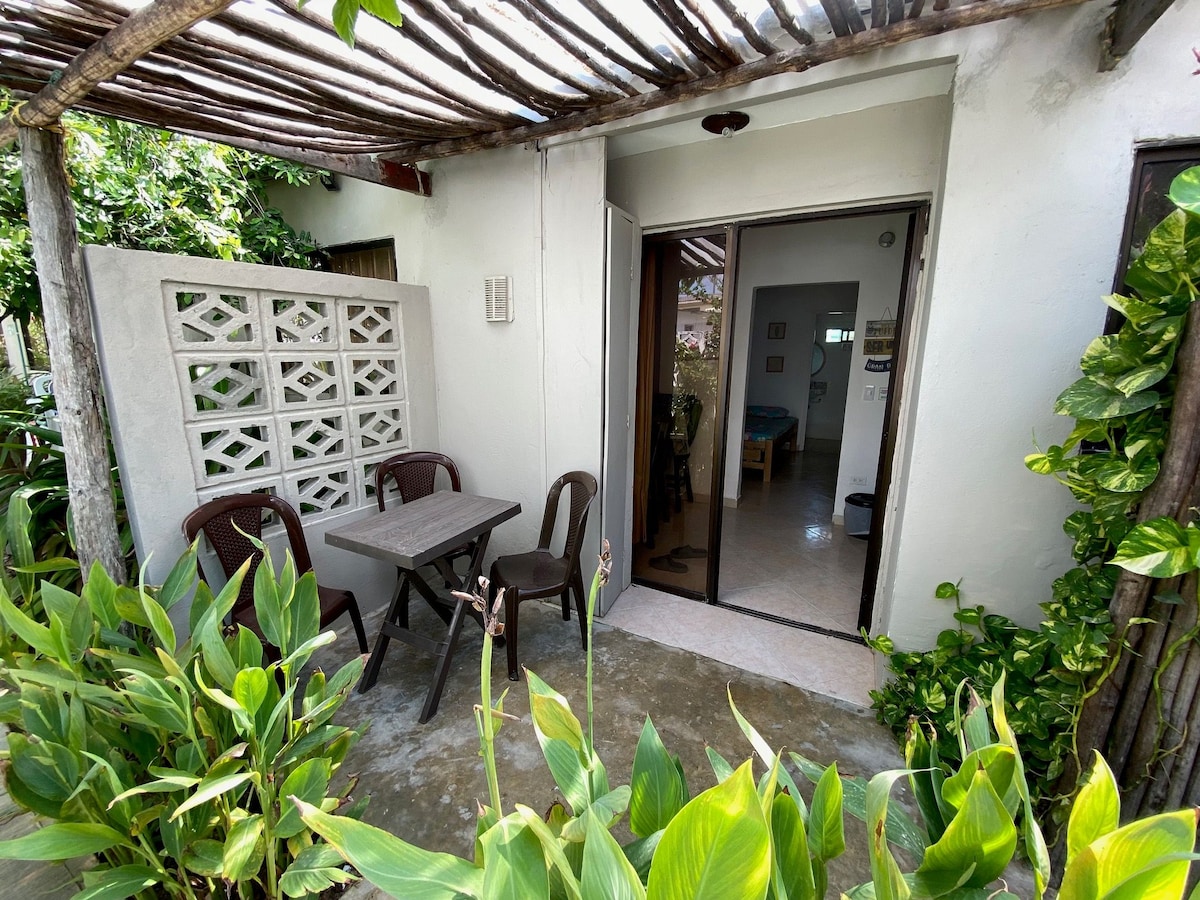 An outdoor seating area is seen, featuring a small table surrounded by two chairs. Vibrant greenery frames the entrance, with a textured wall partially visible. Large sliding glass doors offer a view into the interior space beyond.