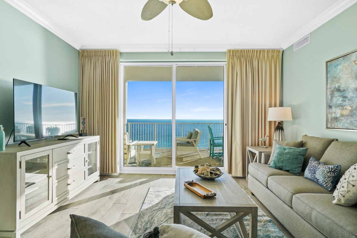 A comfortable living area features light green walls and a neutral sofa adorned with decorative pillows. A coffee table holds a decorative tray, while a large flat-screen TV is positioned against a sideboard. Floor-to-ceiling sliding glass doors provide unobstructed views of the Gulf beyond.