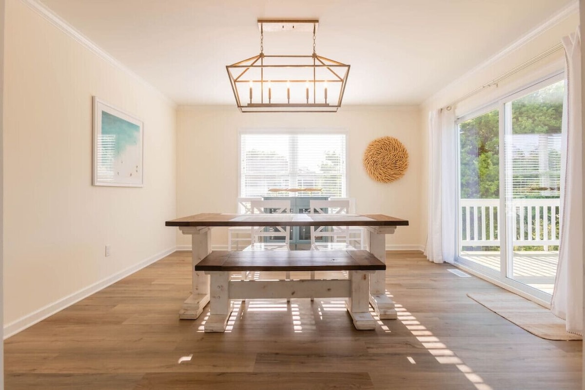 A bright dining area features a large wooden table with a stylish bench. Natural light floods the space through large sliding glass doors, offering views of the outdoor patio. Simple coastal decor adorns the walls, creating a serene environment for meals and gatherings.