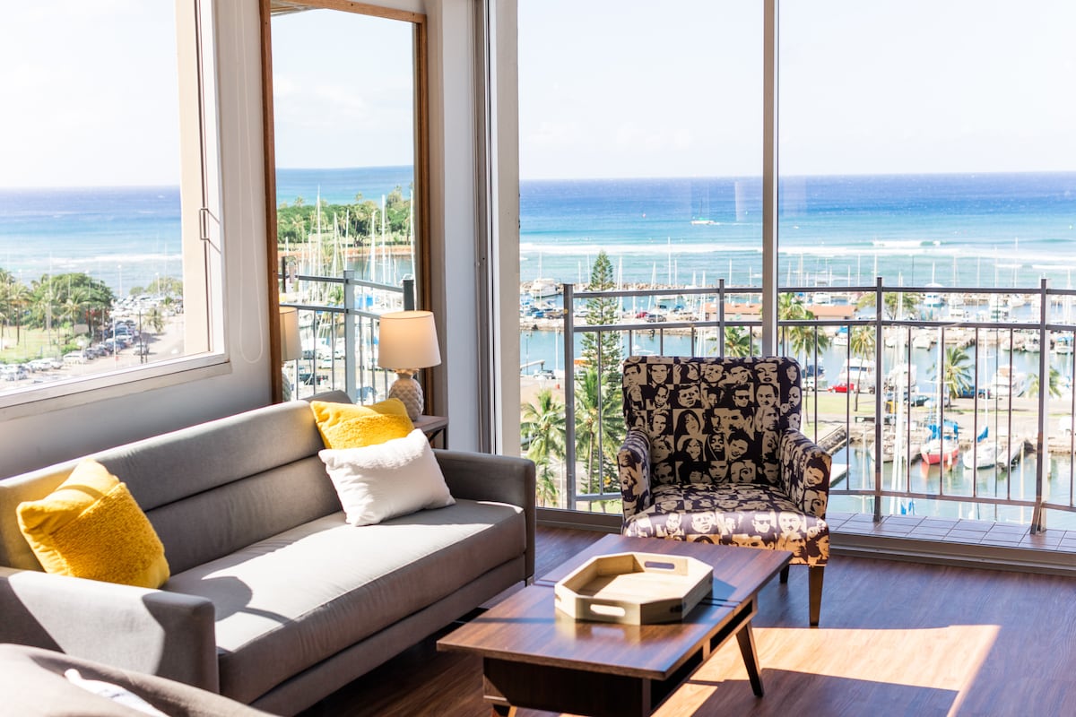 A bright living area features large glass windows offering expansive views of the ocean and marina. A comfortable gray sofa is complemented by a patterned armchair and a wooden coffee table, creating a welcoming spot for relaxation.