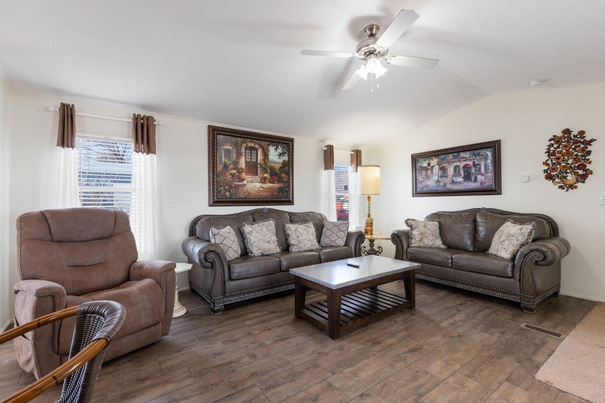 A cozy living area features a pair of plush sofas adorned with decorative pillows, complemented by a recliner chair. A wooden coffee table is centrally placed, with artwork displayed on the walls. Natural light enters through the windows, enhancing the welcoming ambiance.
