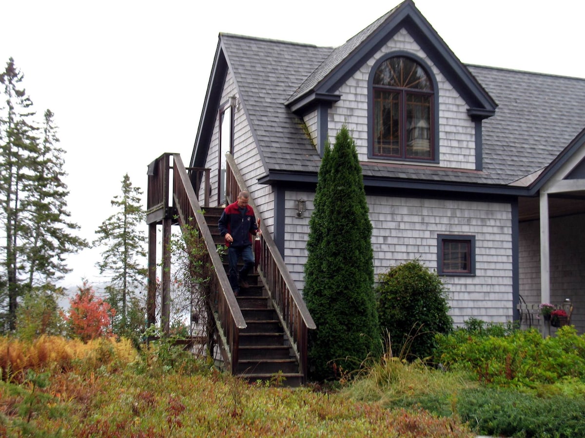 A two-story house is seen with gray shingle siding and a large arched window. A wooden staircase leads to the entrance, surrounded by lush greenery and fall foliage. Evergreen trees frame the property, enhancing the natural setting.