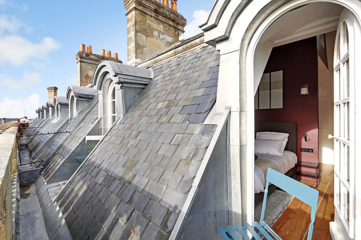 A rooftop balcony offers views of distinctive slate roofing, extending towards arched windows that connect to an inviting interior sleeping area. The room features soft colors and a warm atmosphere, while a simple blue chair is positioned to enjoy the surroundings.