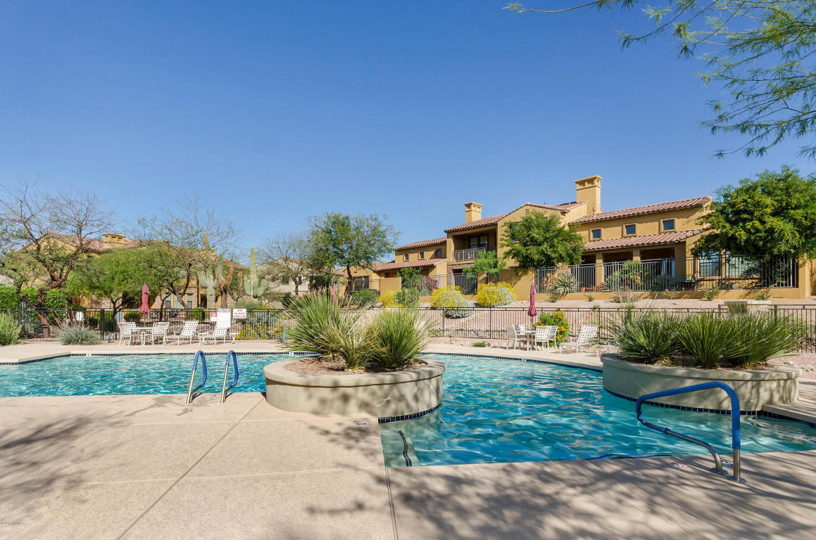 The image displays a swimming pool area surrounded by desert landscape. The pool features a spa-like section with steps leading into the water. Lounge chairs are arranged nearby under the shade of trees. In the background, townhome buildings are visible, complemented by vibrant greenery.