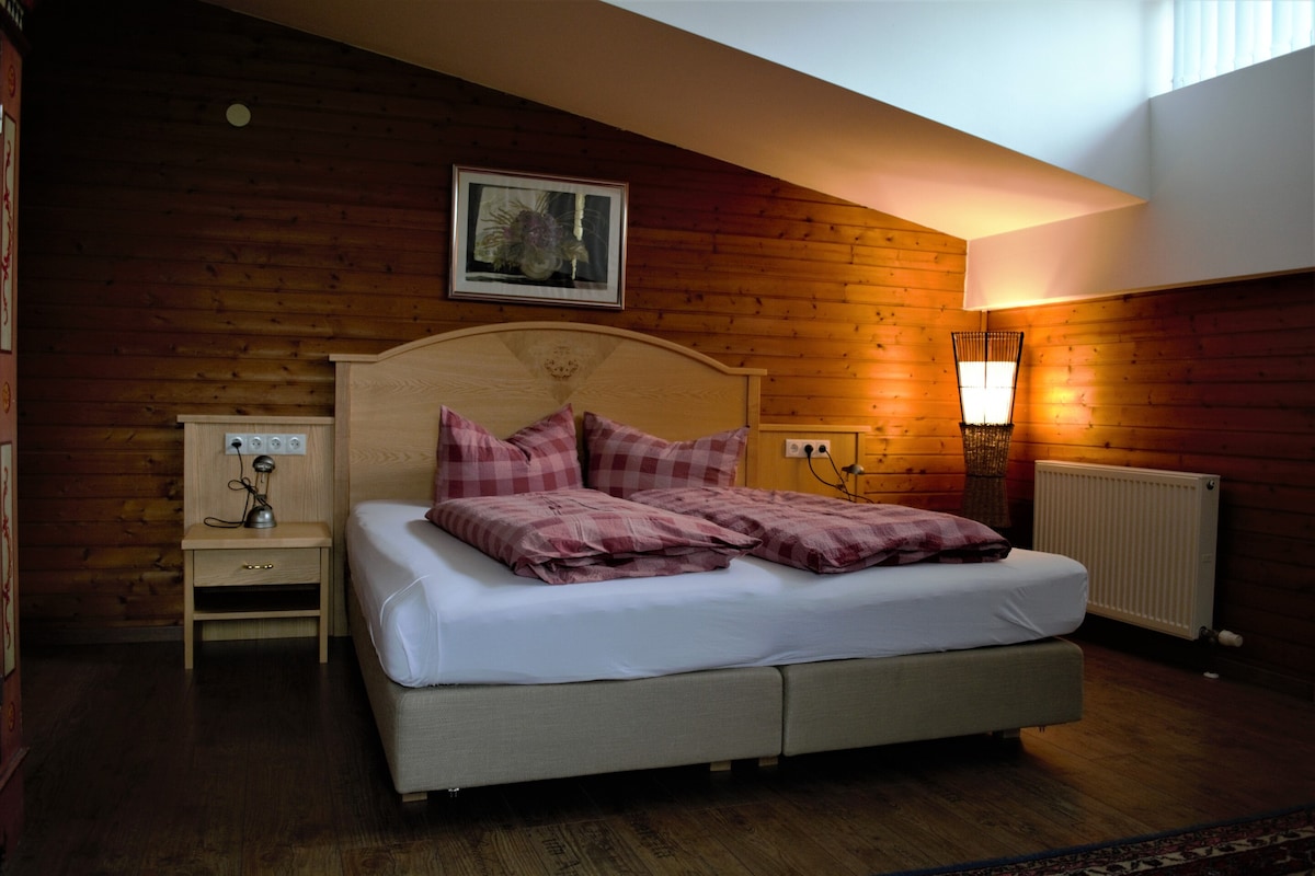 A comfortable sleeping area is featured, with a spacious bed adorned with checkered pillows. Warm wooden walls provide a cozy backdrop, complemented by soft lighting from a standing lamp. A nightstand with a lamp and outlets is positioned beside the bed, enhancing the room's functionality.