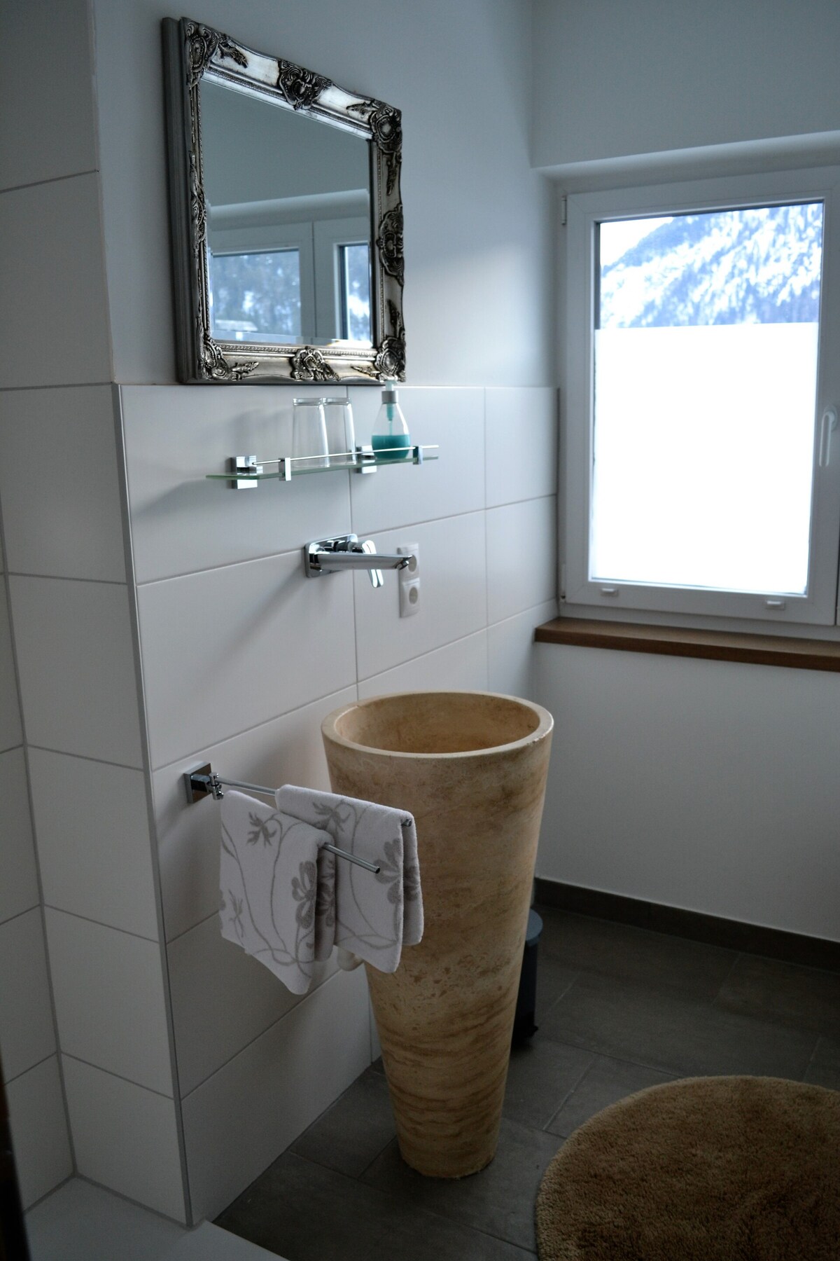 A modern bathroom features a unique stone sink atop a neutral tile surface. A rectangular mirror reflects light from the window, which offers a view of the surrounding mountains. An elegant towel bar holds a neatly folded towel, with a cozy bath mat positioned on the floor.