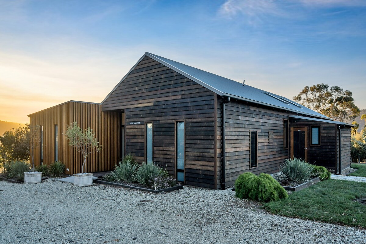 A contemporary farmhouse exterior is presented, showcasing a combination of timber cladding and a modern design. Clean lines highlight large windows that allow natural light to filter in. Surrounding greenery and stone pathways enhance the inviting entrance to Villa Rochford.