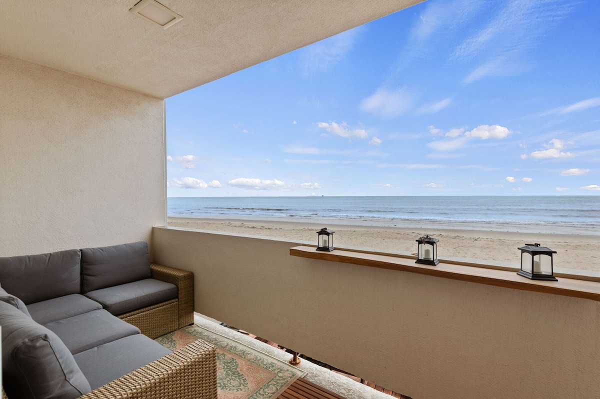 A private outdoor patio provides unobstructed ocean views, featuring a comfortable seating area with grey cushions. The space is enhanced by decorative lanterns placed along the ledge, and a patterned rug adds a touch of warmth to the setting.