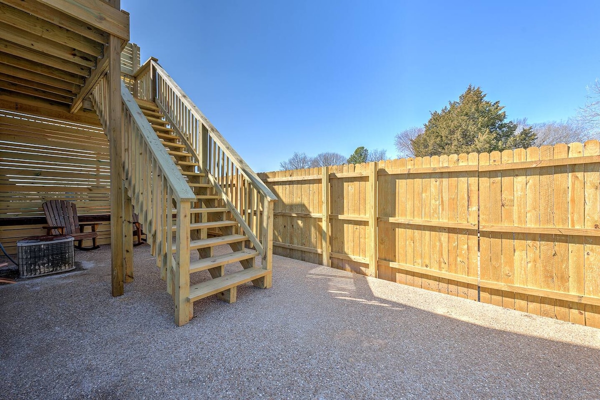 A wooden staircase leads to an elevated deck area, surrounded by a privacy fence. Gravel covers the ground, with two wooden chairs situated near a fire pit in the corner. Bright sunlight illuminates the outdoor space, creating a warm and inviting environment.