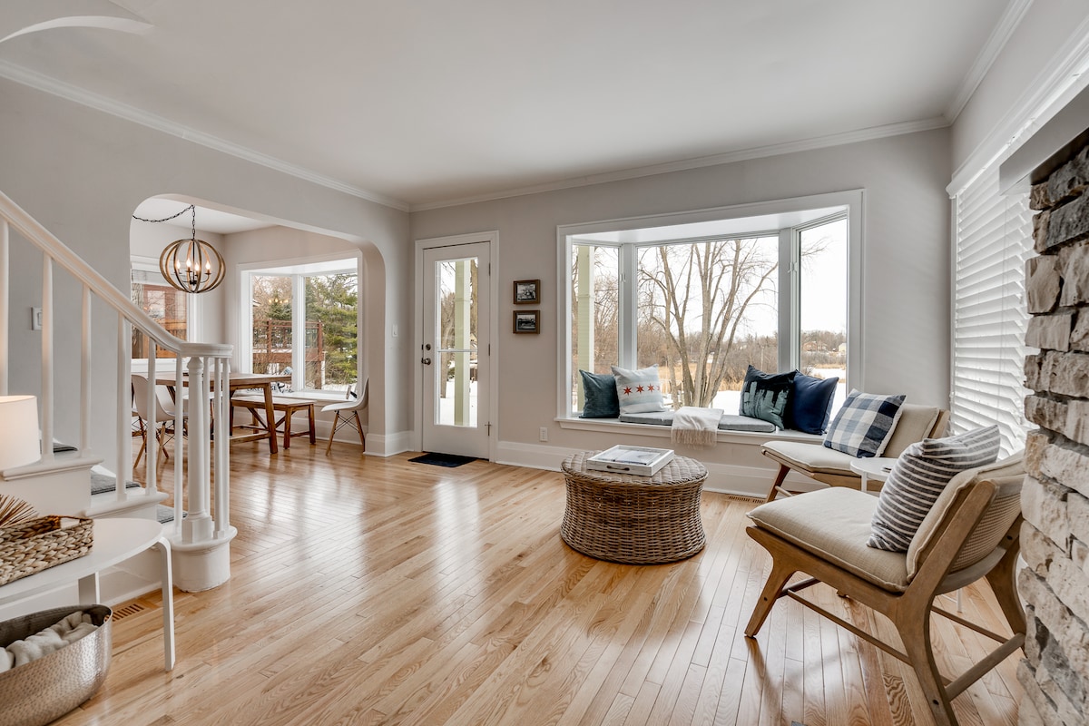 A bright living room is showcased with large windows overlooking outdoor scenery. A cozy seating area includes a light-colored chair and a woven coffee table. Warm wood flooring enhances the spacious feel, complemented by subtle decor and a staircase leading to another level.