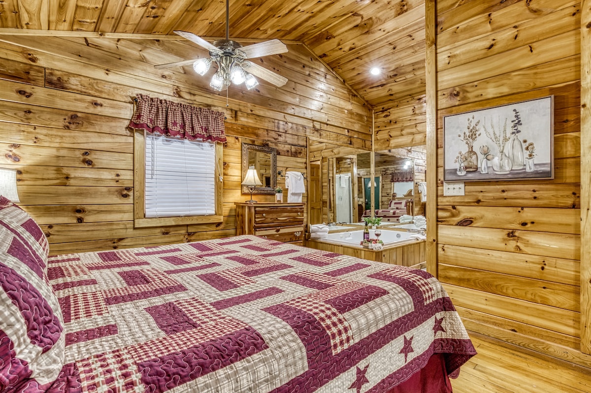 The cabin bedroom features rustic wooden walls and ceiling, creating a warm atmosphere. A spacious bed with a patterned quilt is centered in the room. A wooden dresser and mirror complement the decor, and soft lighting enhances the cozy feel. A window allows for natural light.