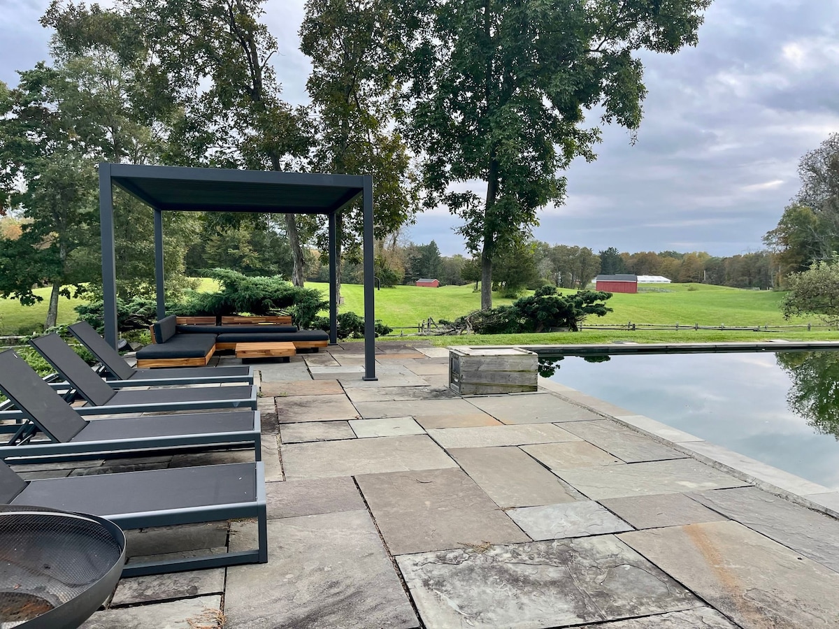 A serene outdoor area features a stone patio with reclining lounge chairs positioned near the edge of a heated pool. A shaded seating structure provides a relaxing space to enjoy the surrounding open fields and distant buildings.