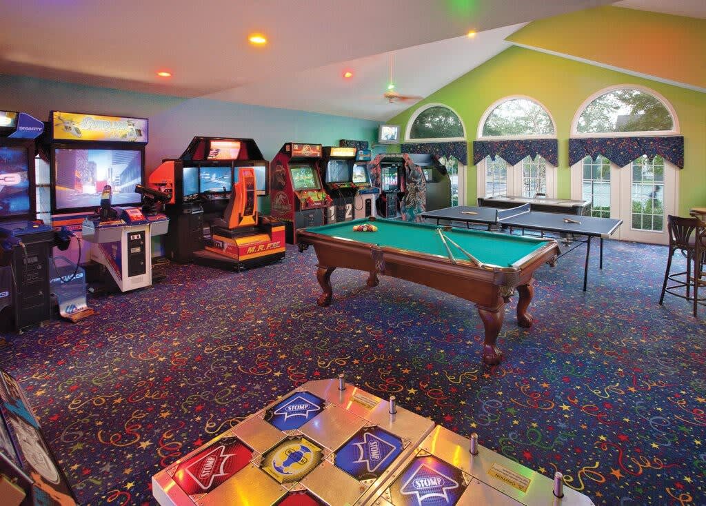 An entertainment area features a variety of arcade game machines and a pool table. Colorful carpet adds a festive touch, while large windows allow natural light to fill the space. The walls are painted in cheerful colors, enhancing the playful environment.