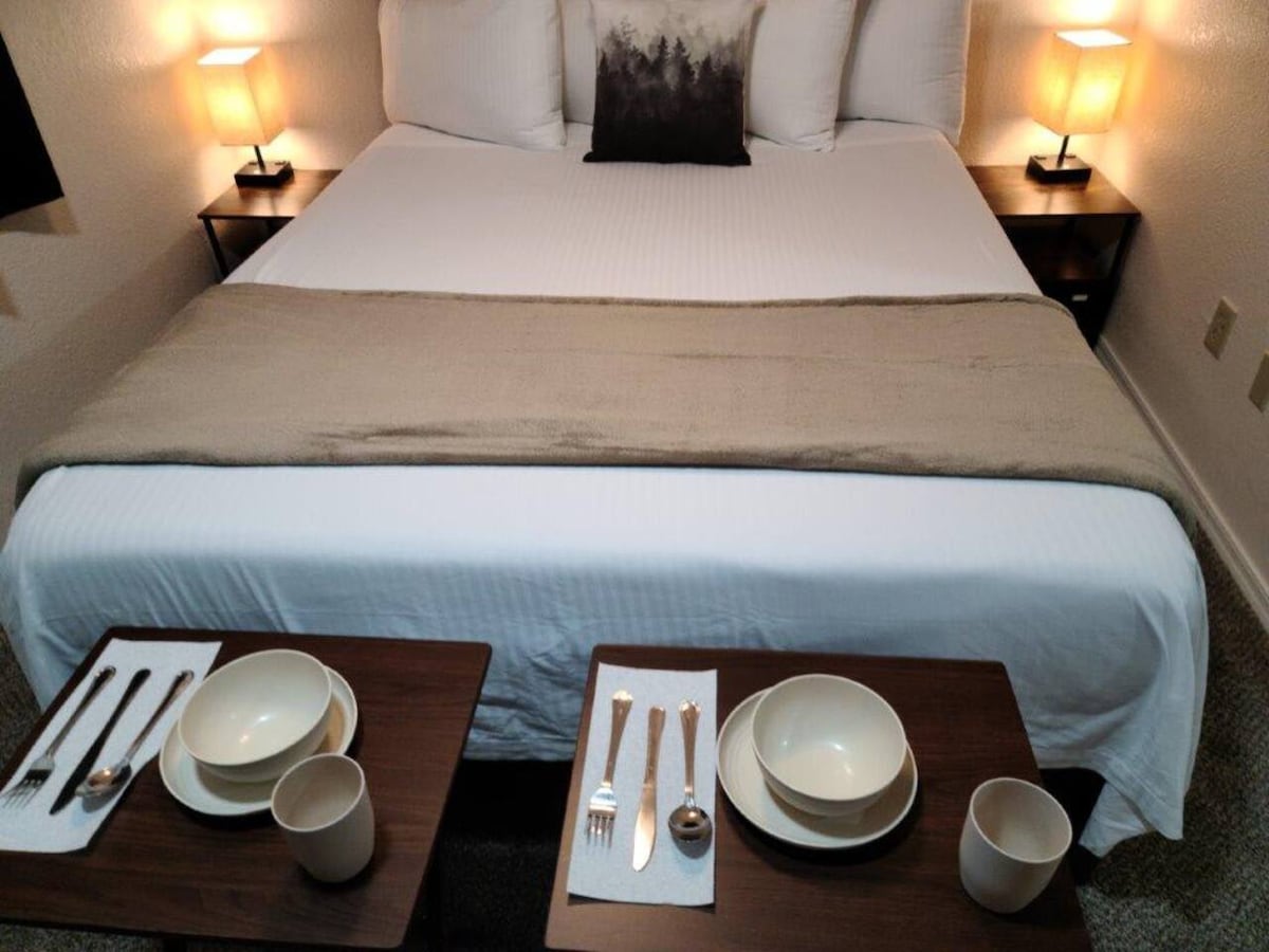 A spacious bed is centered in the image, adorned with a neutral-colored throw and a decorative pillow. Two small side tables hold matching bowls and cups, alongside silverware neatly arranged on placemats. Soft lighting from bedside lamps creates a warm ambience.