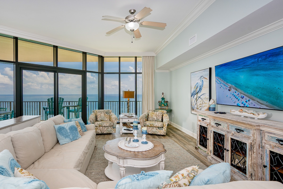 The living area features a large sectional sofa arranged around a circular coffee table. A flat-screen television is mounted on the wall, with calming coastal artwork nearby. Floor-to-ceiling windows offer expansive views of the Gulf, while furnishings and decor reflect a coastal aesthetic.