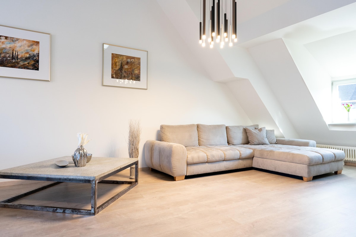 A spacious living area is highlighted by a light-colored sectional sofa positioned against a sloped ceiling. A modern coffee table sits centrally, accompanied by decorative elements. Natural light streams in through a nearby window, enhancing the room's airy feel.
