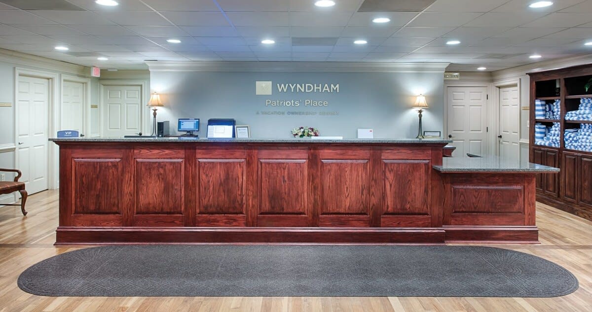 The reception area features a polished wooden front desk, complemented by soft lighting and decorative plants. A dark area rug lies beneath the desk, enhancing the overall welcoming ambiance. The backdrop includes neatly organized shelves and framed signage indicating Wyndham Patriots Place.
