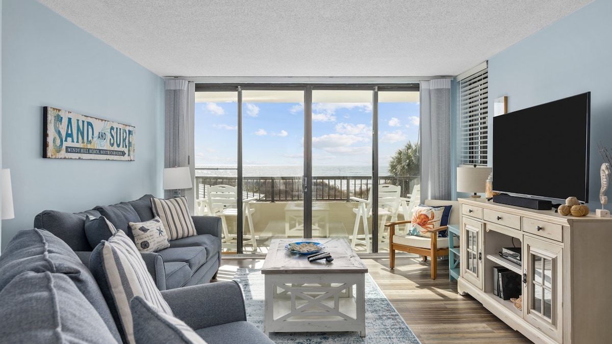 A cozy living area features a light blue wall and plush seating arranged for relaxation. Large glass doors provide access to a balcony with ocean views. A flat screen TV is positioned on a light wood console, complemented by a decorative coffee table and nautical accents.