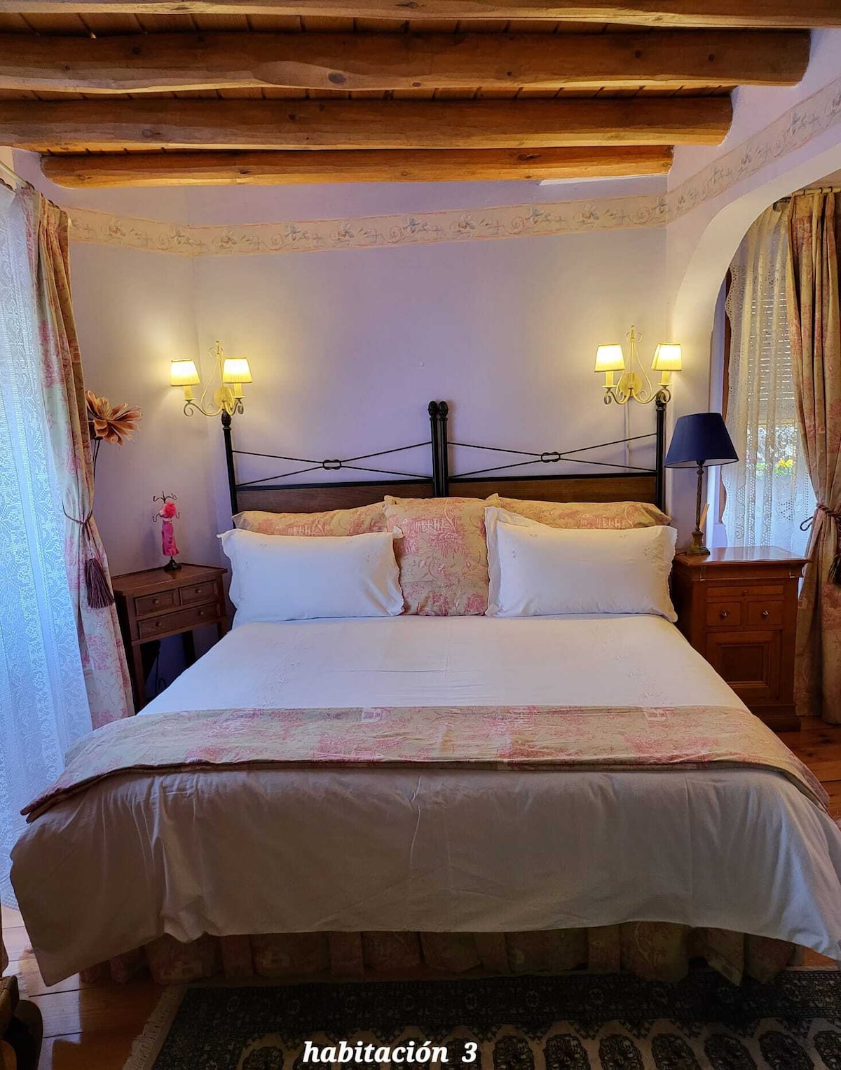 A cozy double bedroom features a spacious bed dressed with soft linens and decorative pillows. Two bedside tables frame the bed, with lamps providing gentle light. Elegant curtains adorn the window, and rustic wooden beams are visible on the ceiling, creating a warm ambiance.