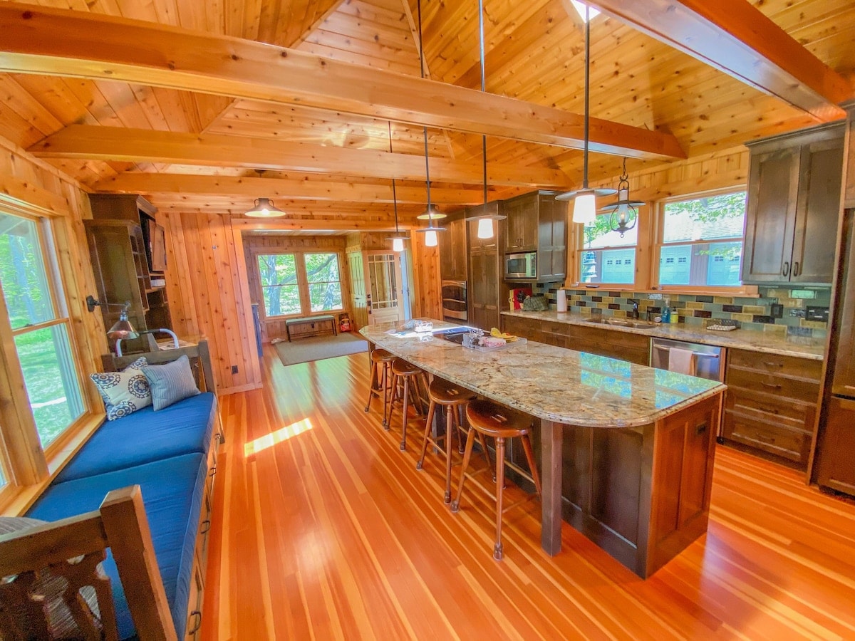 A spacious kitchen and living area feature wooden beams and warm wooden accents. The kitchen island is equipped with seating, and natural light flows through large windows, illuminating the space. A cozy nook with a blue cushion invites relaxation.