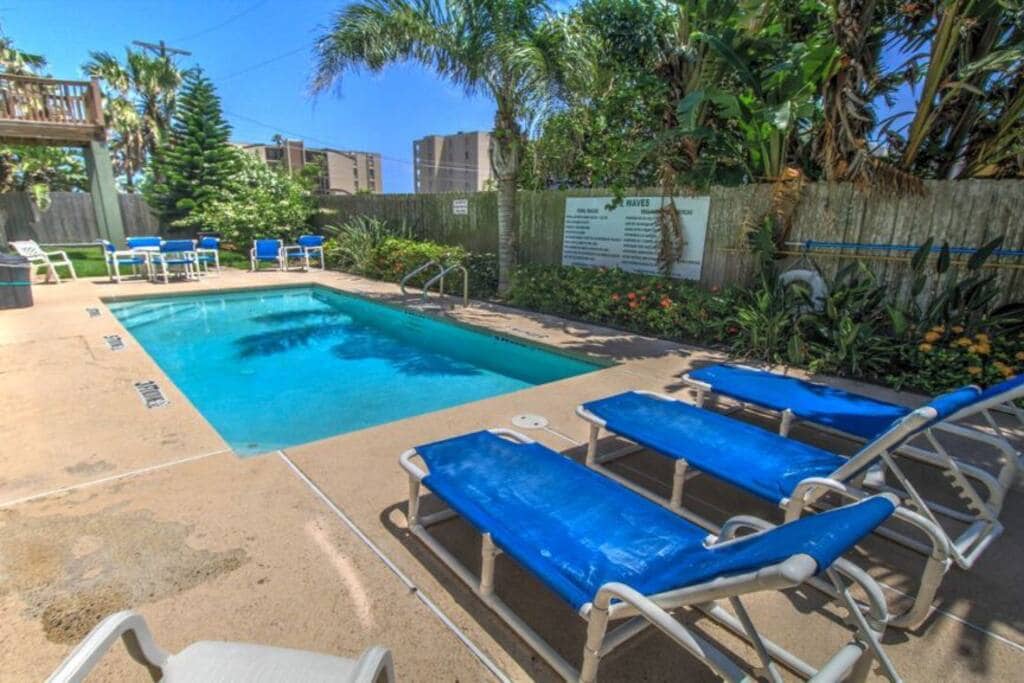A tranquil pool area is surrounded by lush tropical landscaping. Blue lounge chairs are positioned around the pool, offering relaxation space. The clear water of the pool reflects the bright blue sky, while palm trees provide shade along the edges.