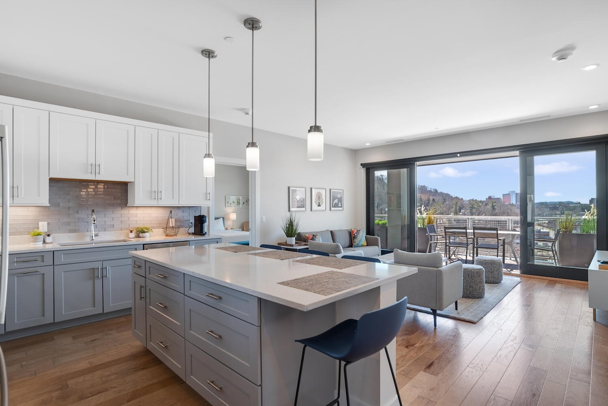 A contemporary kitchen features a central island with seating and sleek cabinetry. Bright pendant lights hang above, illuminating the space. Large sliding doors open to a balcony showcasing views of the mountains. Natural light floods the area, enhancing the open floor plan.
