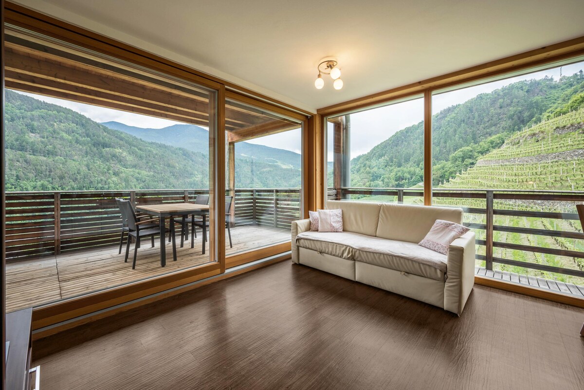 A spacious living area is captured with large glass doors that provide a view of the surrounding mountains and greenery. A comfortable white sofa is positioned in front of the doors, while a dining table and chairs are visible on the balcony.