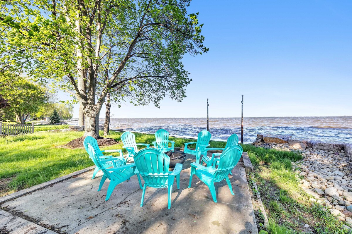 A spacious outdoor area is set up with turquoise chairs arranged around a fire pit. The background features a scenic view of the water, framed by trees and a gentle shoreline.