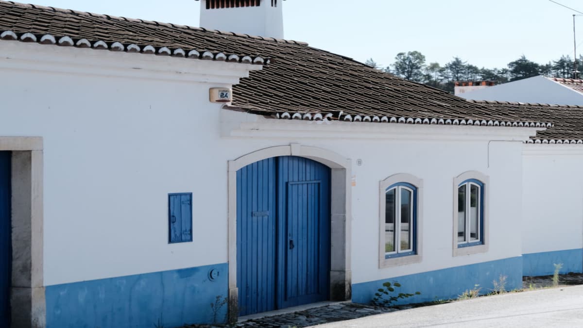 The exterior of a charming white cottage is depicted, featuring a blue door and matching window frames. The sloped roof showcases traditional tiles, contributing to the building's rustic charm. A small window on the left adds to the overall character of the structure, which sits along a quiet street.