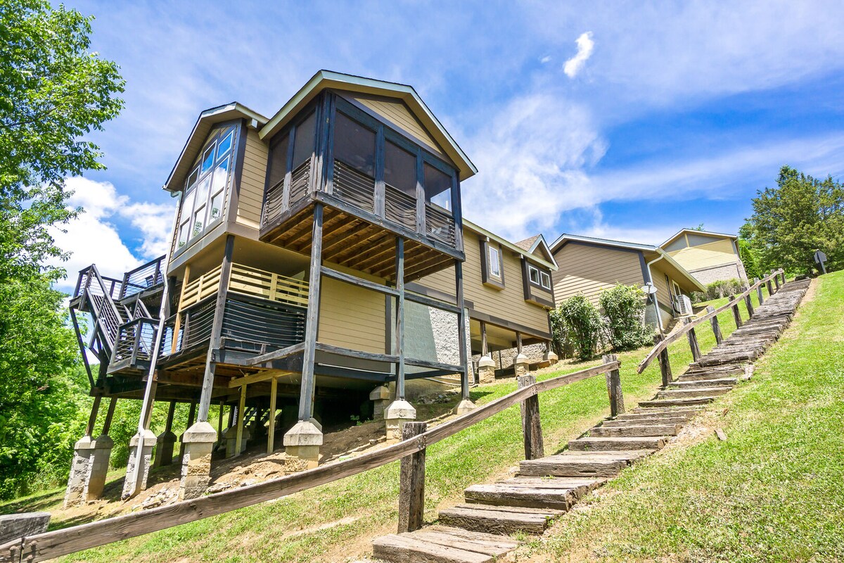 A two-story house is set on a hillside, supported by sturdy stilts. The exterior features a mix of light and dark colors, with large windows allowing for natural light. A wooden staircase leads up the grassy slope, flanked by trees and landscaped areas.