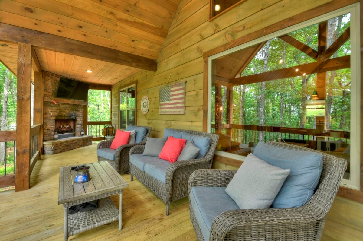 A spacious back porch is furnished with comfortable seating, featuring two light blue sofas with coral accent pillows and a coffee table. Natural greenery surrounds the area, creating a peaceful outdoor setting. A wood-burning fireplace can be seen in the background, adding to the ambiance.