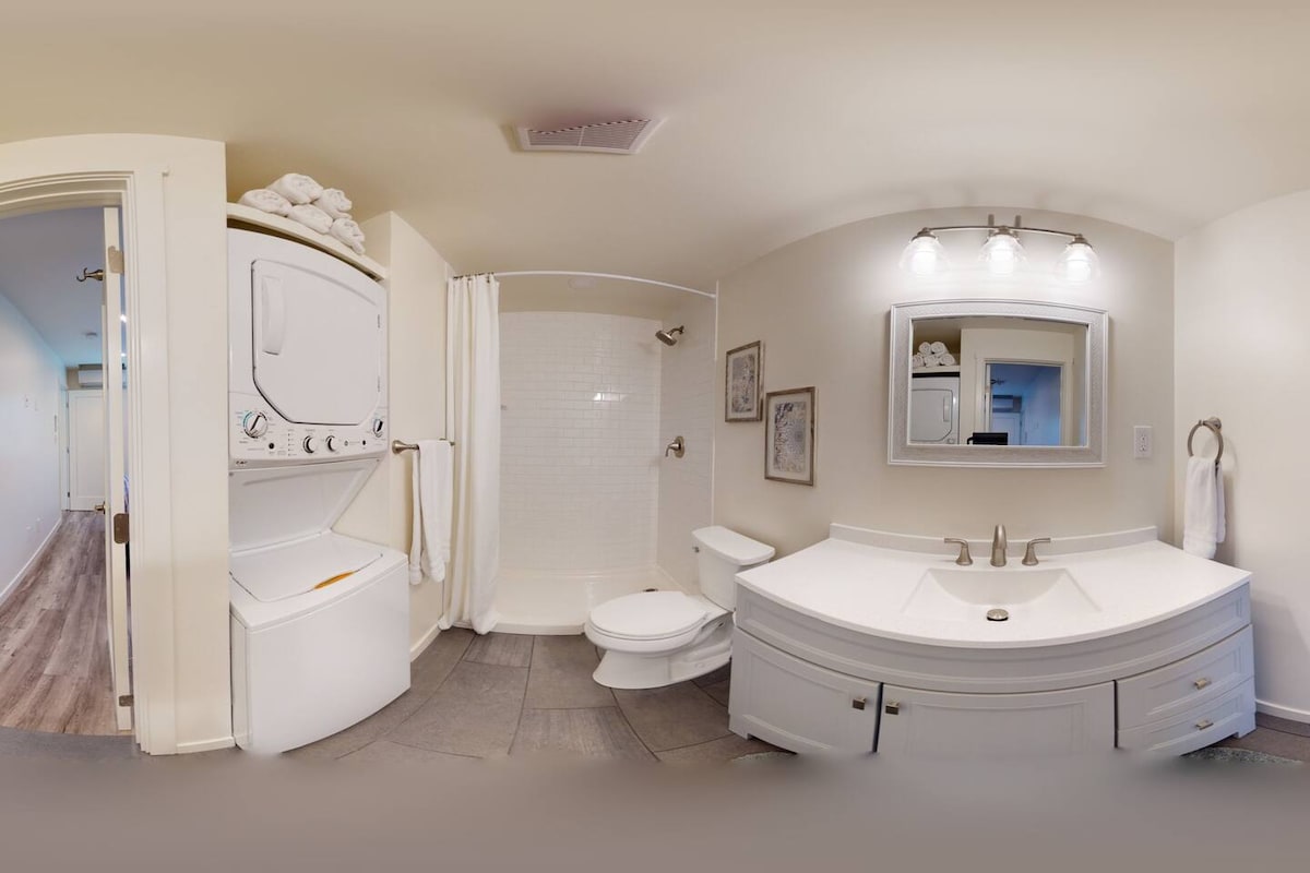 A modern bathroom features a spacious vanity with dual sinks and a large mirror above. A shower area with a glass door is visible along with a toilet. A stacked washer and dryer unit is conveniently placed in the corner, with white towels neatly arranged nearby.
