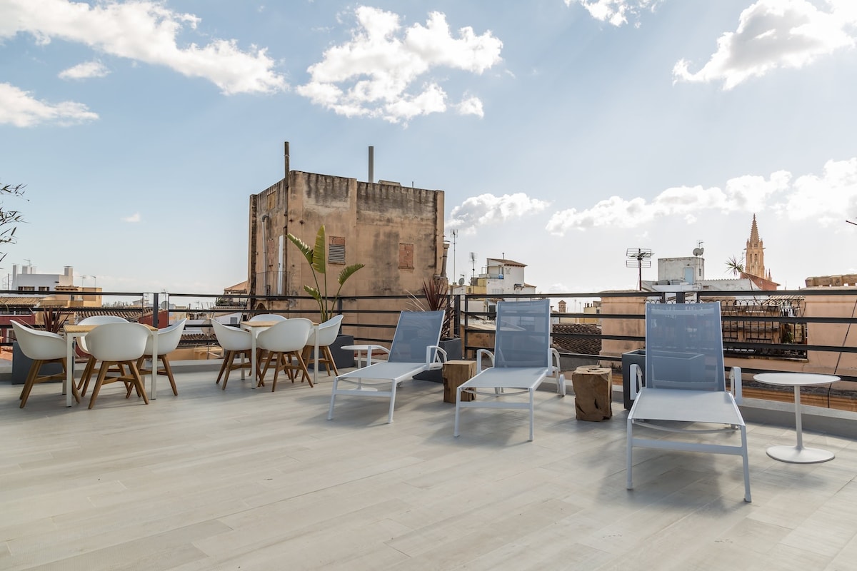 A rooftop terrace features a blend of lounge chairs and dining tables arranged for relaxation and socializing. The backdrop includes urban structures and a glimpse of a church steeple, under a bright sky with scattered clouds.
