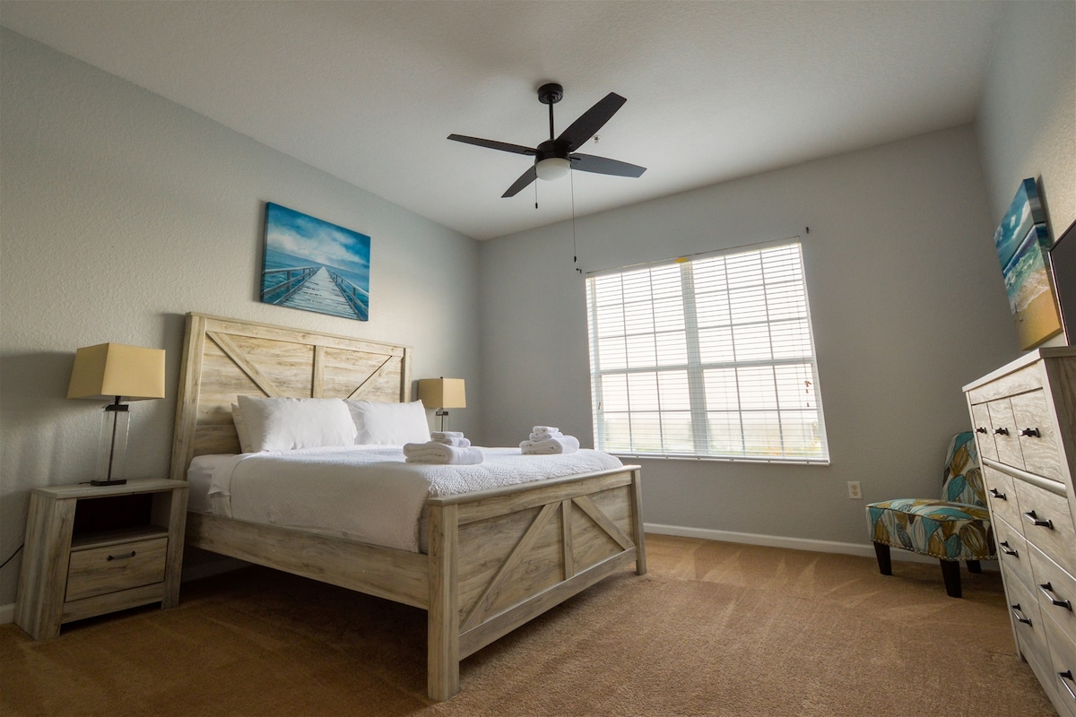 A well-lit bedroom features a spacious bed with freshly laundered linens. A ceiling fan hangs above, and light filters through the window adorned with blinds. A stylish chair is positioned by the window, complemented by a wooden dresser and a vibrant piece of wall art.