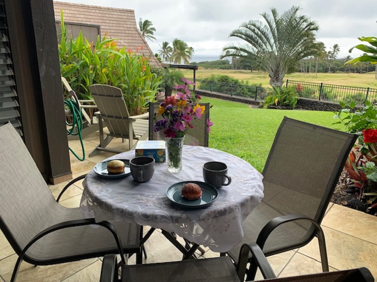 A small round table is set for breakfast on a covered lanai, featuring two plates with pastries and two cups. A vibrant floral arrangement adds color, while the green lawn and palm trees are visible in the background, creating a relaxed outdoor dining space.