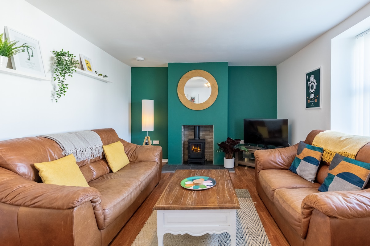 The lounge features two leather sofas, with decorative cushions arranged comfortably. A coffee table sits at the center, and a smart TV is positioned nearby. A traditional stove with a gas fire adds a cozy element, complemented by a round mirror above the fireplace.