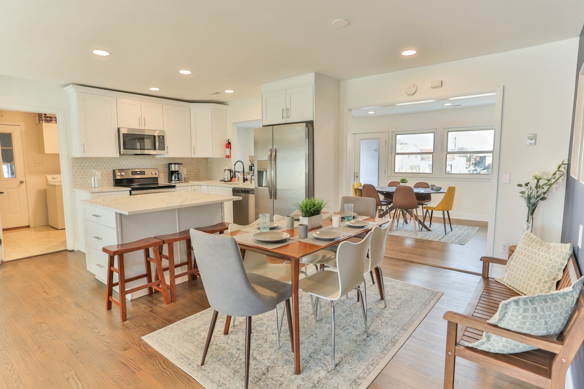 A modern kitchen is visible, featuring white shaker cabinets, quartz countertops, and stainless steel appliances. A central island with seating complements a dining area with a large table surrounded by colorful chairs. Natural light streams through multiple windows, creating a welcoming atmosphere.