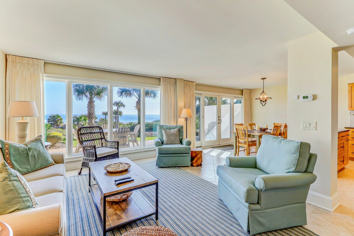 A spacious living area features large windows offering views of the ocean and palm trees. Comfortable seating is arranged around a wooden coffee table. A dining area is visible in the background, illuminated by natural light streaming through the glass doors.