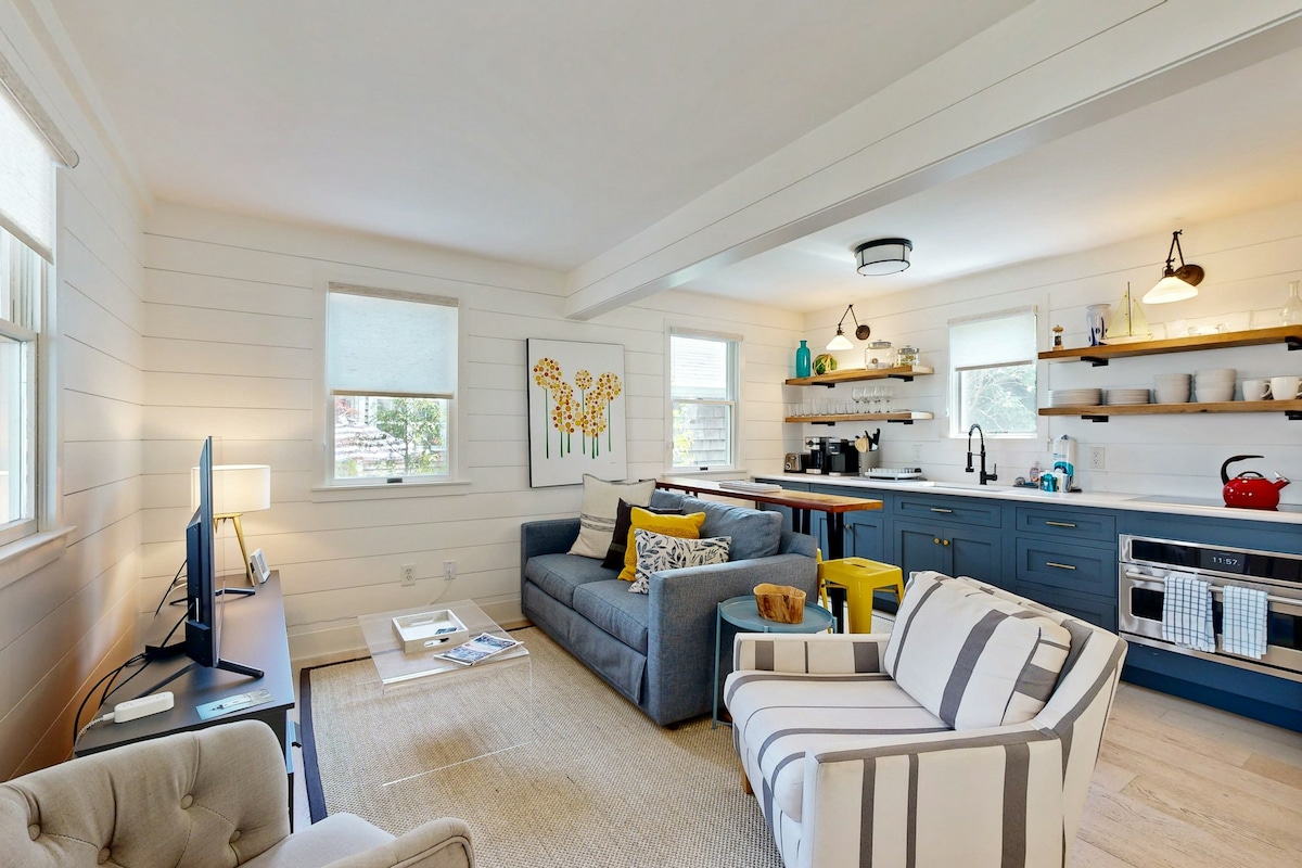 A bright living area features a gray sofa and a striped armchair, inviting relaxation. A television is positioned on a console table. The kitchen showcases blue cabinetry and modern appliances, with a yellow bar stool adding a pop of color. Natural light streams through windows.