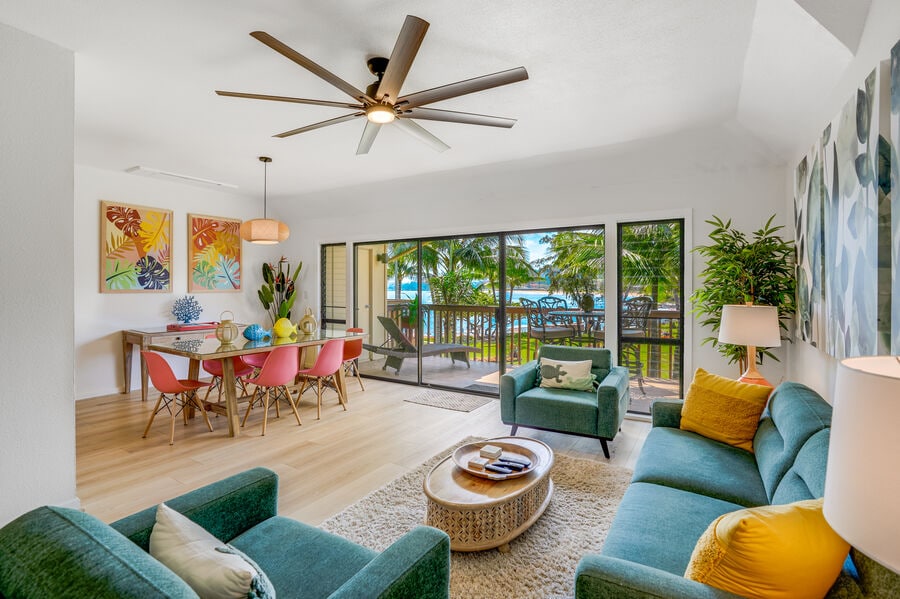An open living area is presented with a comfortable seating arrangement, featuring a plush turquoise sofa and an accompanying chair. A dining table is set for meals, surrounded by colorful chairs. Large glass doors provide access to a balcony with views of palm trees and the ocean.