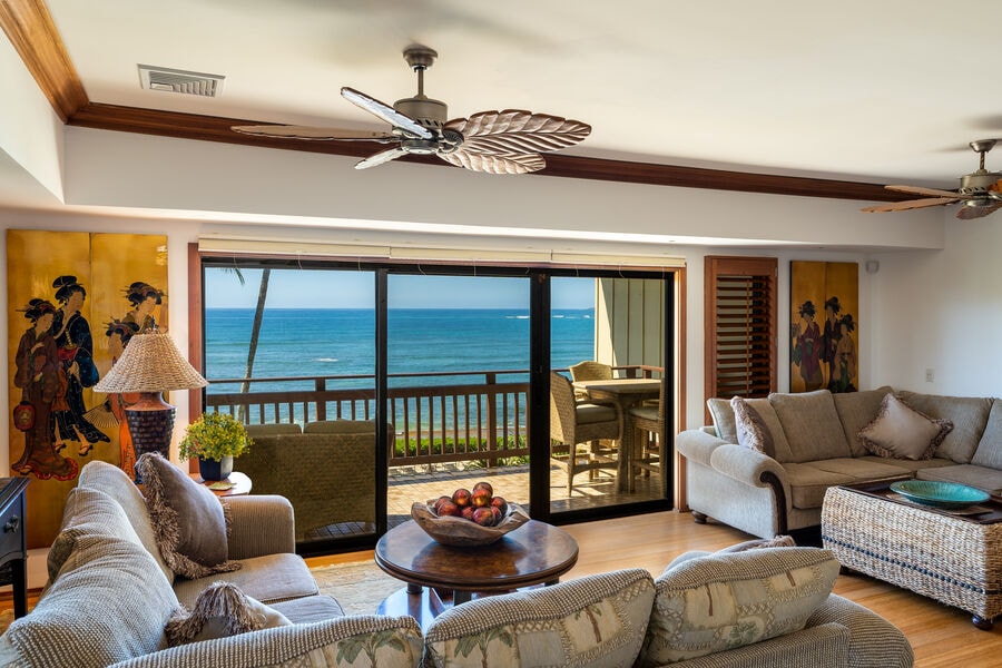 A sunlit living area features large glass doors that open to a scenic ocean view. Two comfortable sofas surround a central coffee table adorned with fresh fruit. Tropical artwork decorates the walls, complementing the coastal atmosphere. Ceiling fans are positioned overhead, providing cooling breeze.