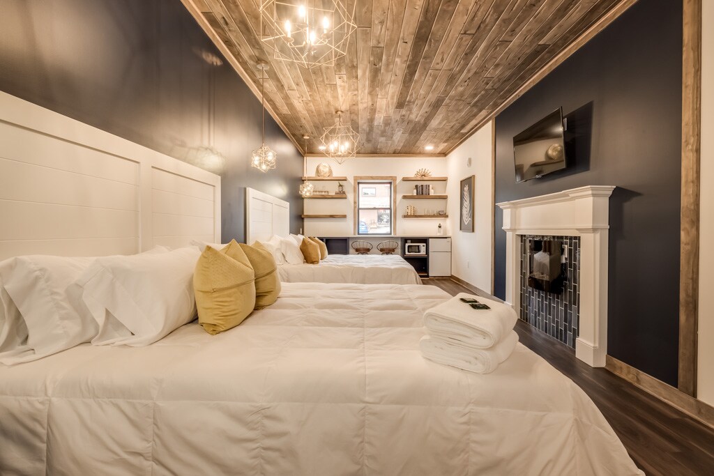 A spacious hotel suite showcases two plush king beds dressed in white bedding, accompanied by yellow accent pillows. A cozy electric fireplace is positioned beneath a mounted television, complemented by sleek shelving displaying decorative items. Warm wood paneling and contrasting dark walls enhance the elegant ambiance.