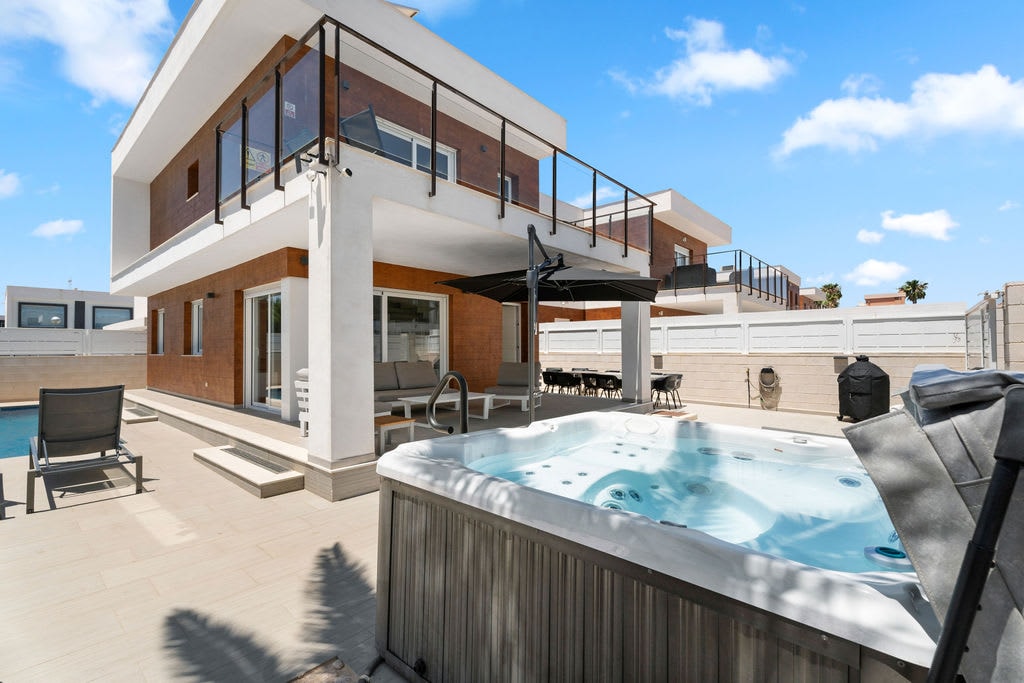 An outdoor space features a private jacuzzi adjacent to a pool area, with modern loungers positioned nearby. The villa is set against a clear blue sky, showcasing a spacious terrace area with a dining setup. A shaded seating area is also visible.