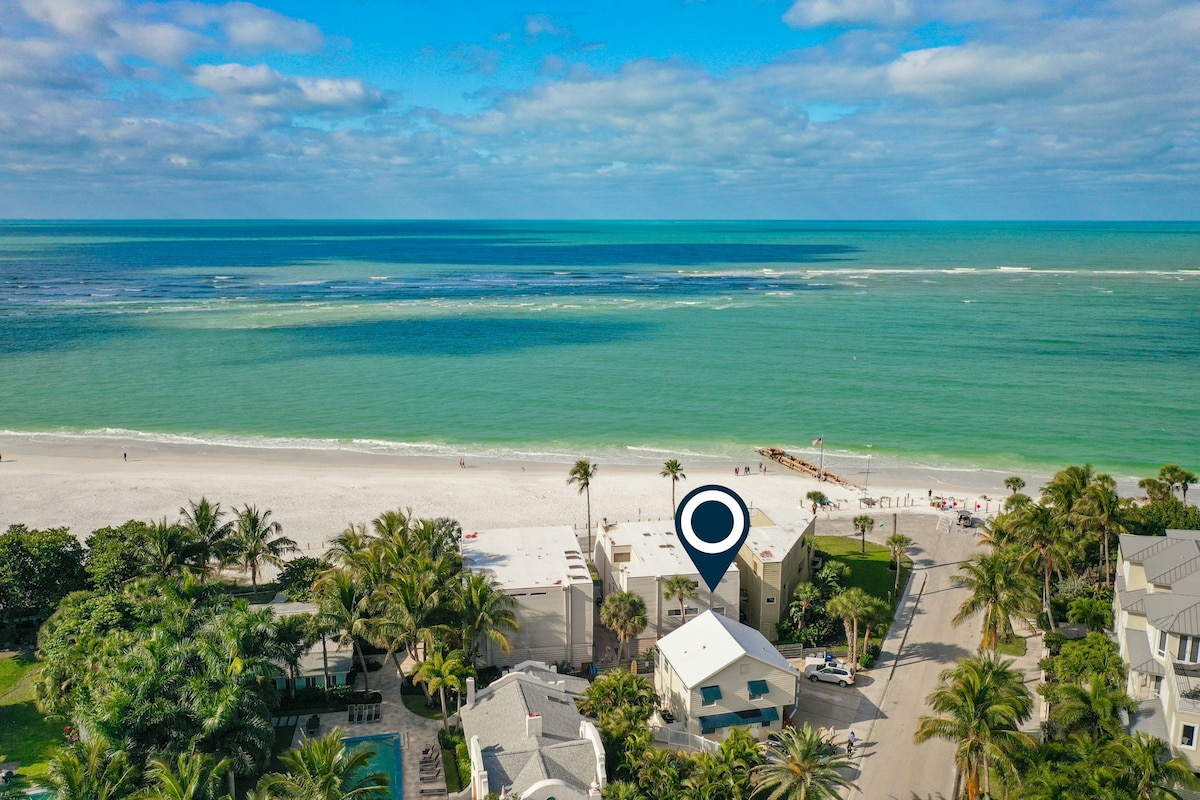 An aerial view captures the proximity of Parrot Beach Cottages to the shoreline, with bright blue waters meeting soft white sands. Surrounding palm trees offer a tropical ambiance, while beach access and a nearby pier are visible in the background, enhancing the coastal setting.