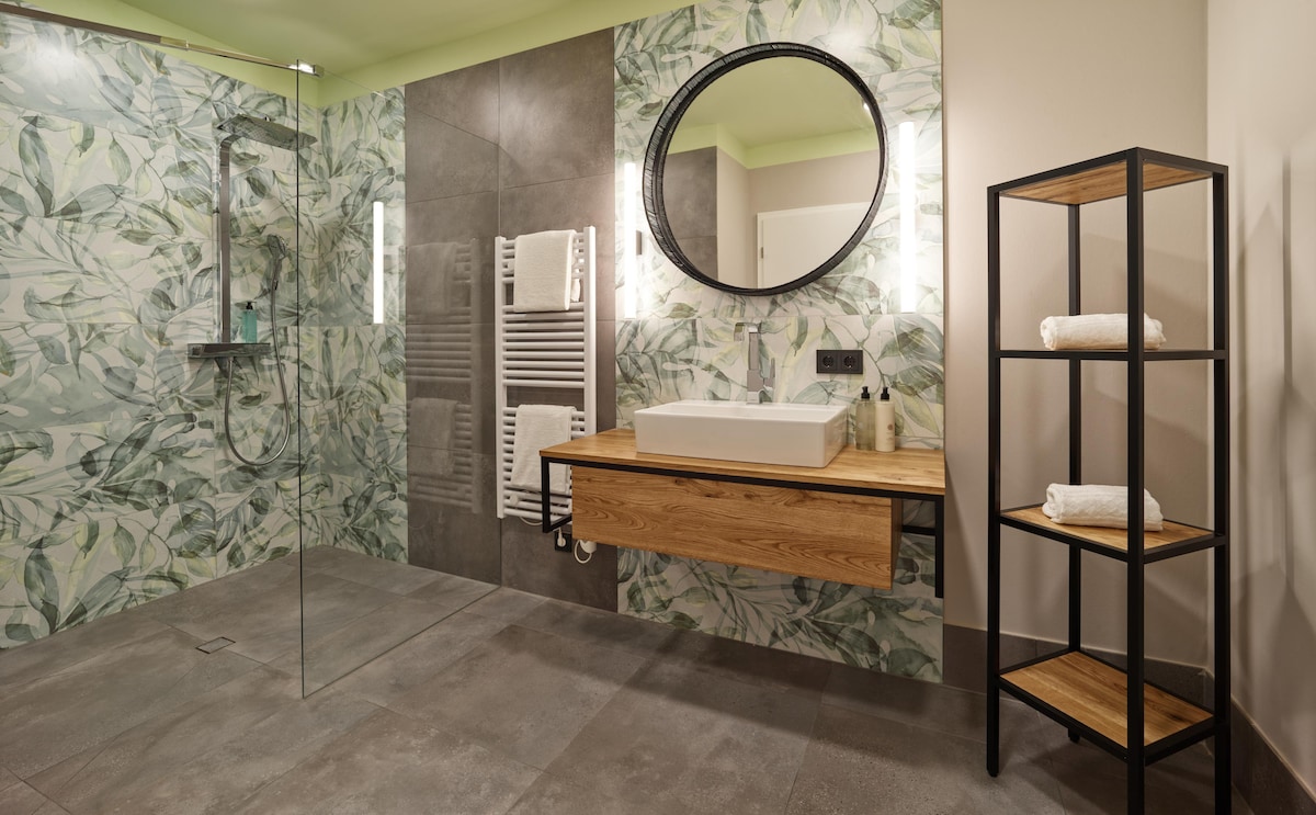A modern bathroom features a spacious walk-in shower with glass panels and a tropical leaf-patterned wall. A circular mirror above a wooden vanity complements a minimalist shelf displaying neatly folded white towels.